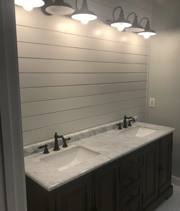 Modern bathroom vanity with dual sinks, marble countertop, dark cabinetry, and industrial-style pendant lighting against shiplap wall.