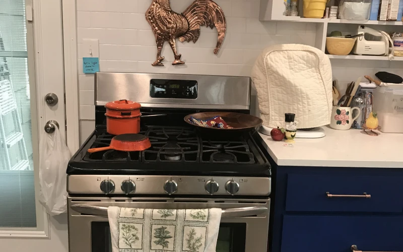 Modern kitchen remodel featuring a stainless steel gas range with copper hood, white subway tile backsplash, decorative copper rooster art, blue cabinetry, open shelving, and herb-patterned tea towel accents.