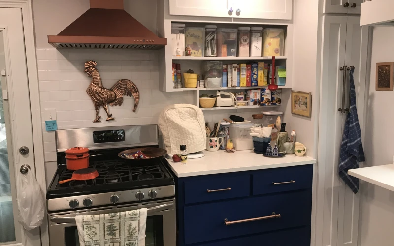Modern kitchen remodel featuring navy blue cabinetry with copper hardware, white countertops, stainless steel gas range with copper range hood, and open shelving with organized storage.