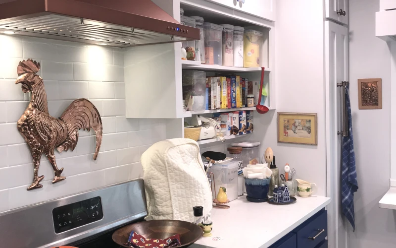 Modern kitchen remodel featuring navy blue cabinetry, white countertops, burgundy range hood, gas cooktop, open shelving, and subway tile backsplash with decorative rooster accent.