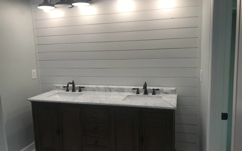 Modern bathroom vanity with dual sinks, marble countertop, dark cabinetry, and shiplap wall accented by black barn-style lighting fixtures.